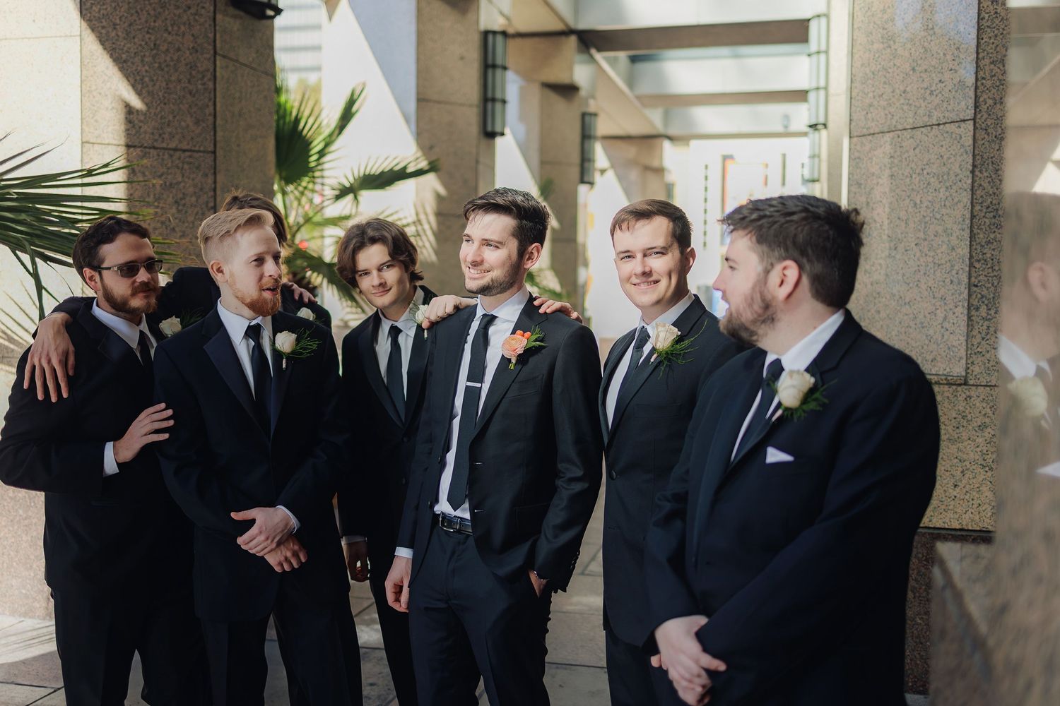 A group of groomsmen in black suits and ties share a candid moment while standing together at a wedding venue.