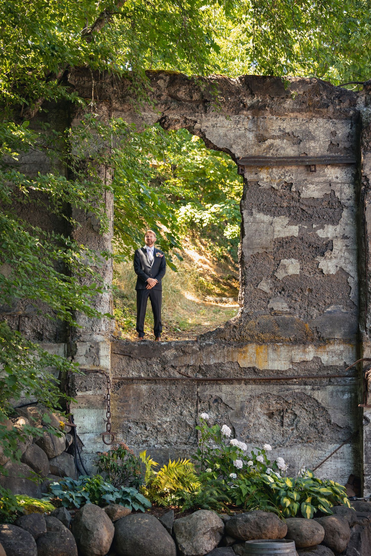A figure stands in a beautiful stone archway surrounded by lush greenery and rocks in a garden setting.