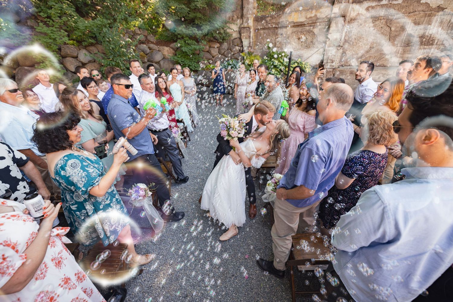 Guests throw confetti at a joyous outdoor celebration while standing along both sides of a stone pathway.
