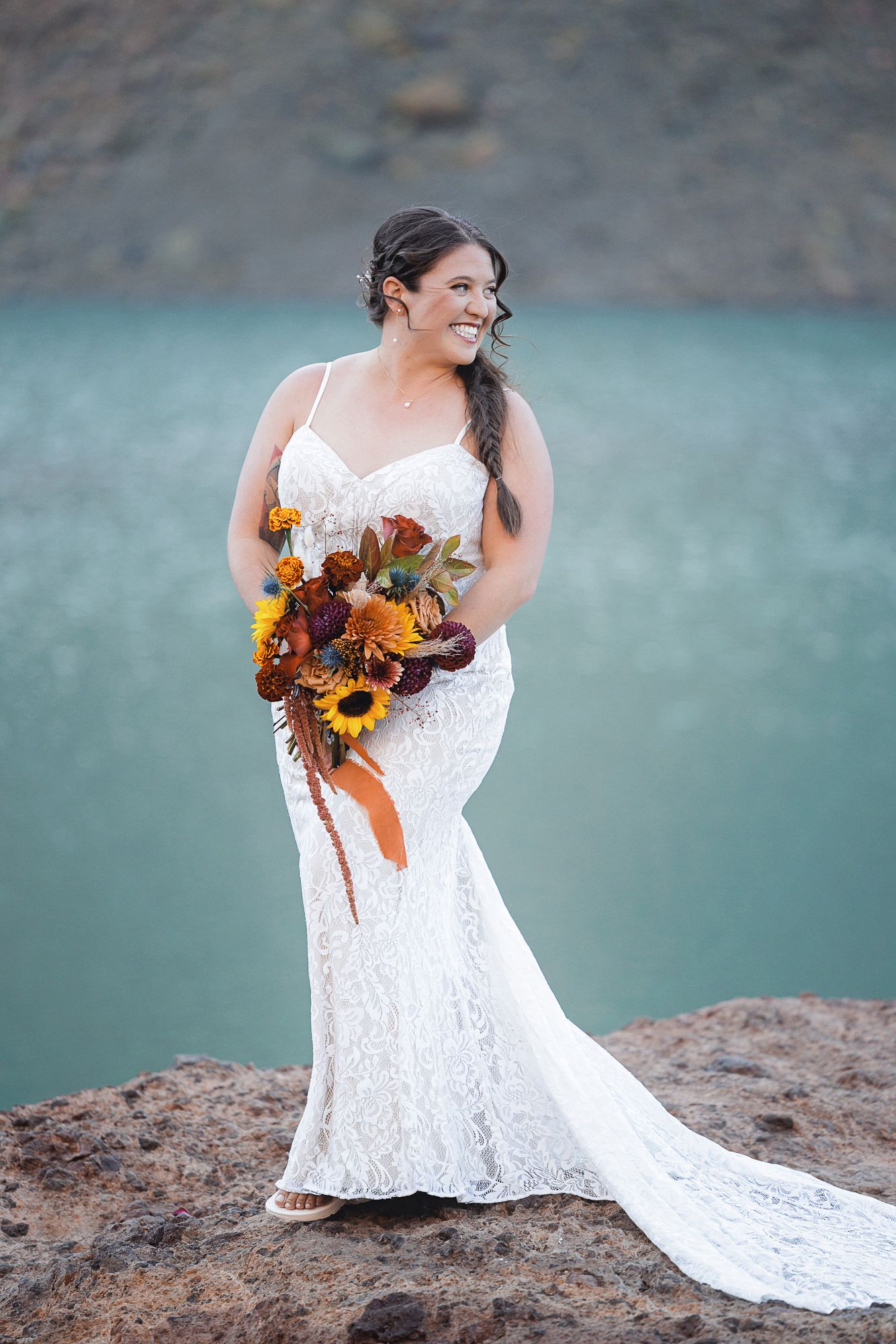 Smiling bride in a lace wedding gown holds an autumn sunflower bouquet while standing on a rocky ledge above a turquoise lake.