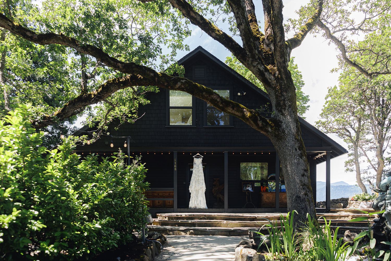 Black craftsman cabin surrounded by lush trees with a white wedding dress hanging on the front porch.