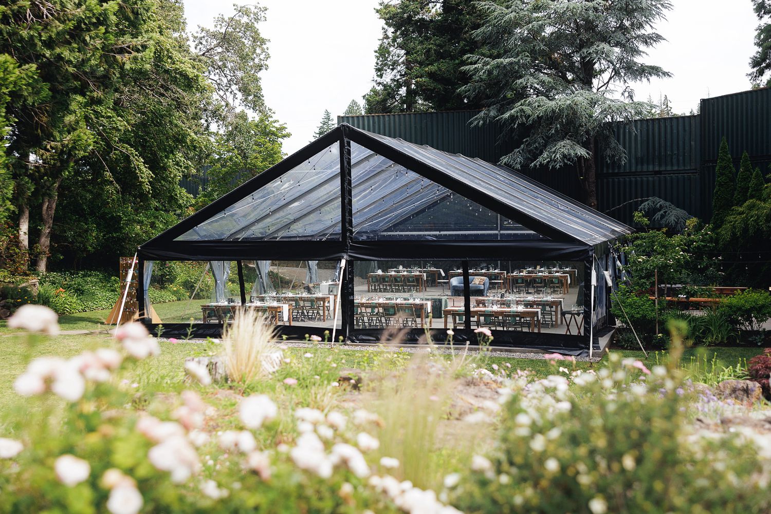 Elegant outdoor marquee tent with clear walls and dark frame, set in a lush garden with wildflowers.