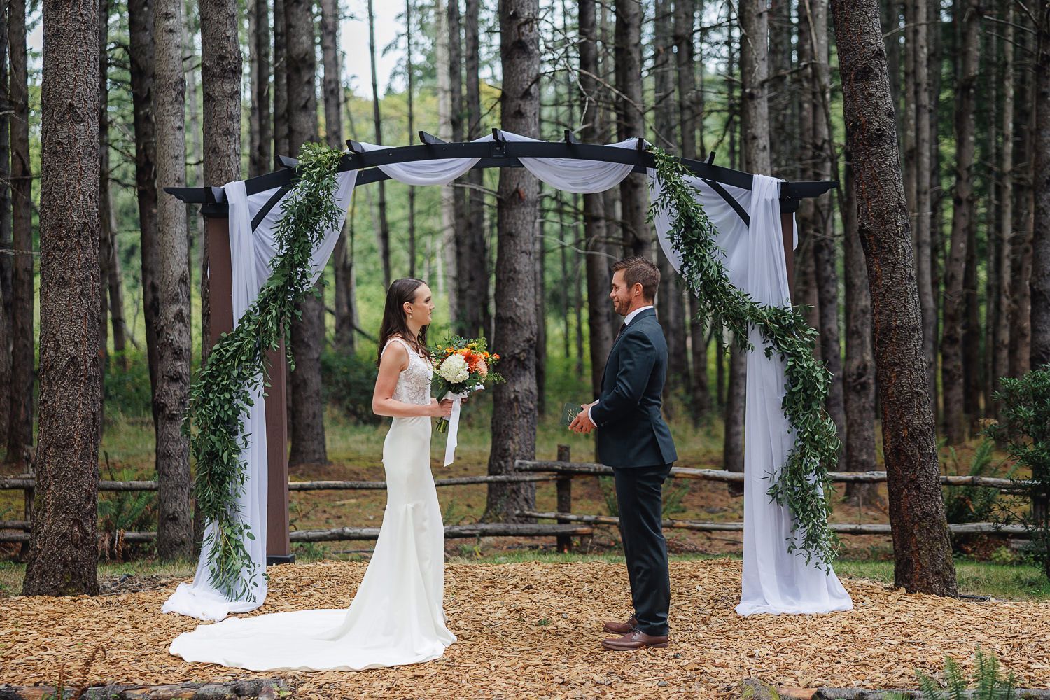 A romantic outdoor wedding ceremony takes place in a serene pine forest with white fabric draped between trees.