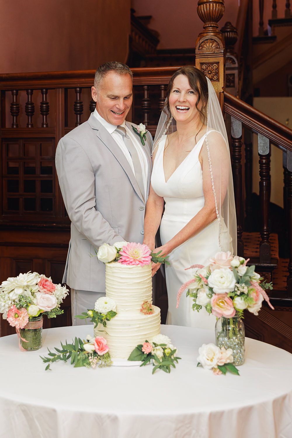 A couple in wedding attire cut their elegant white wedding cake decorated with pink and white roses at a reception.