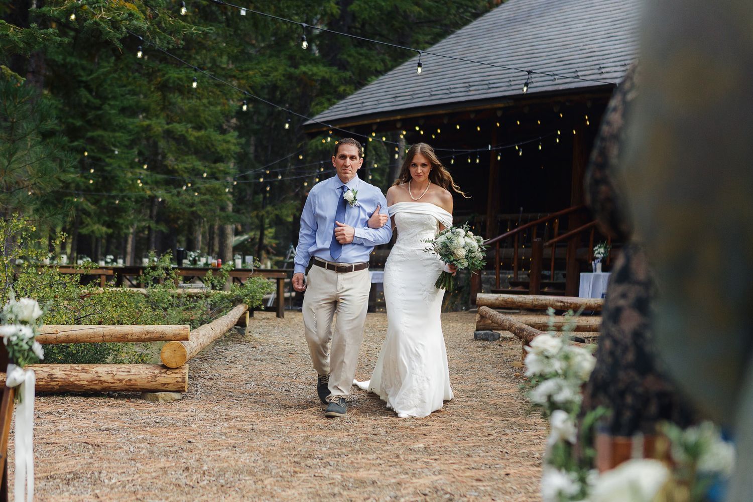 A couple walks together on a path under string lights near a rustic wooden building.