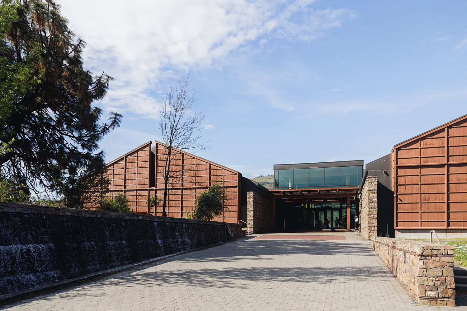Modern brick building with angular rooflines, glass entrance, and paved driveway surrounded by trees under a blue sky.