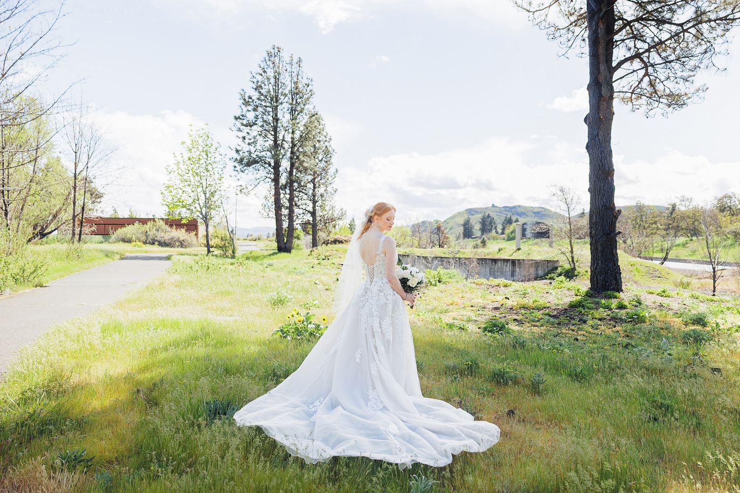 Bride in elegant white gown with long train stands in lush green meadow surrounded by trees on her wedding day.
