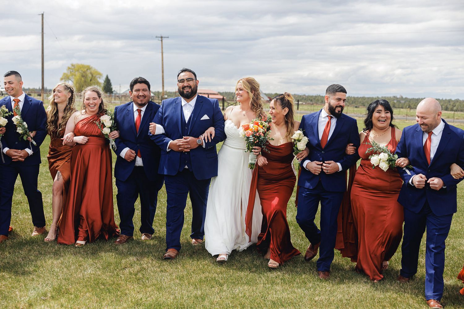 Wedding party in navy suits and rust bridesmaid dresses laughing outdoors on a green lawn.