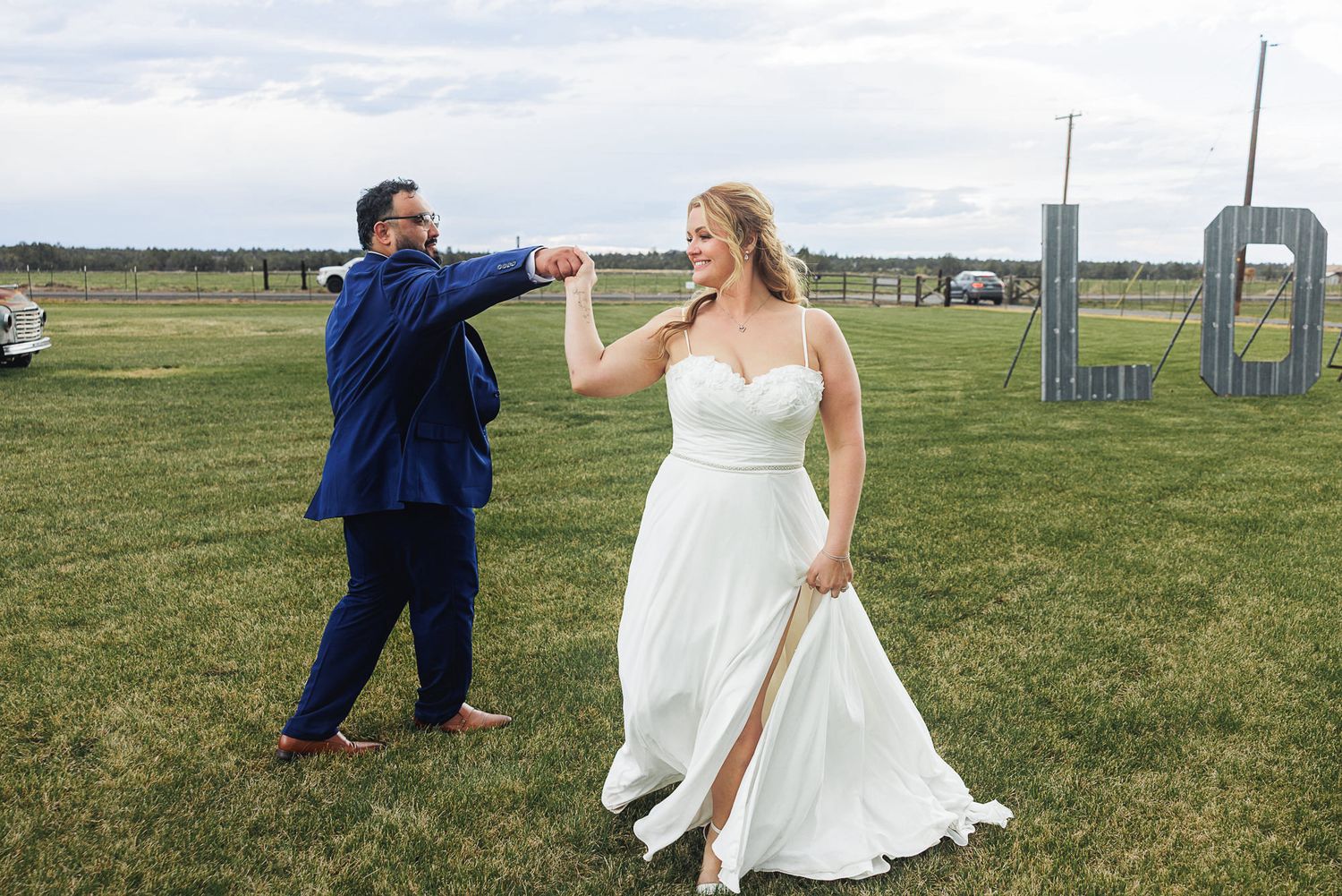 Bride and groom dancing outdoors on green grass at wedding venue, bride in white gown, groom in blue suit.