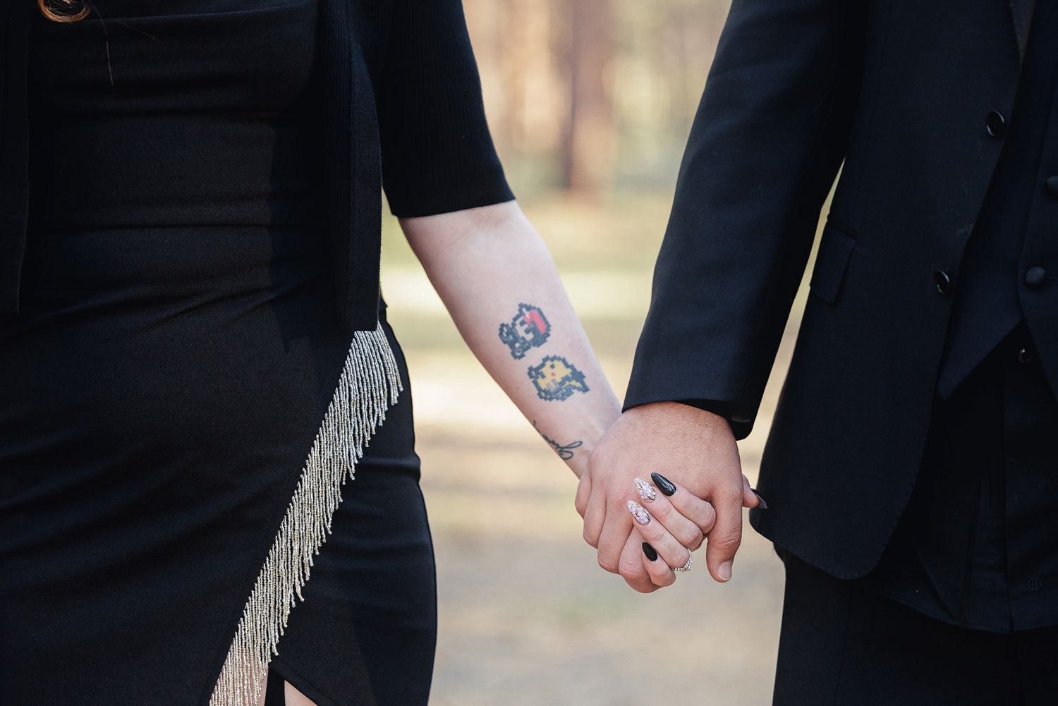 Couple holding hands outdoors, one with colorful tattoos on wrist, both dressed in black attire.