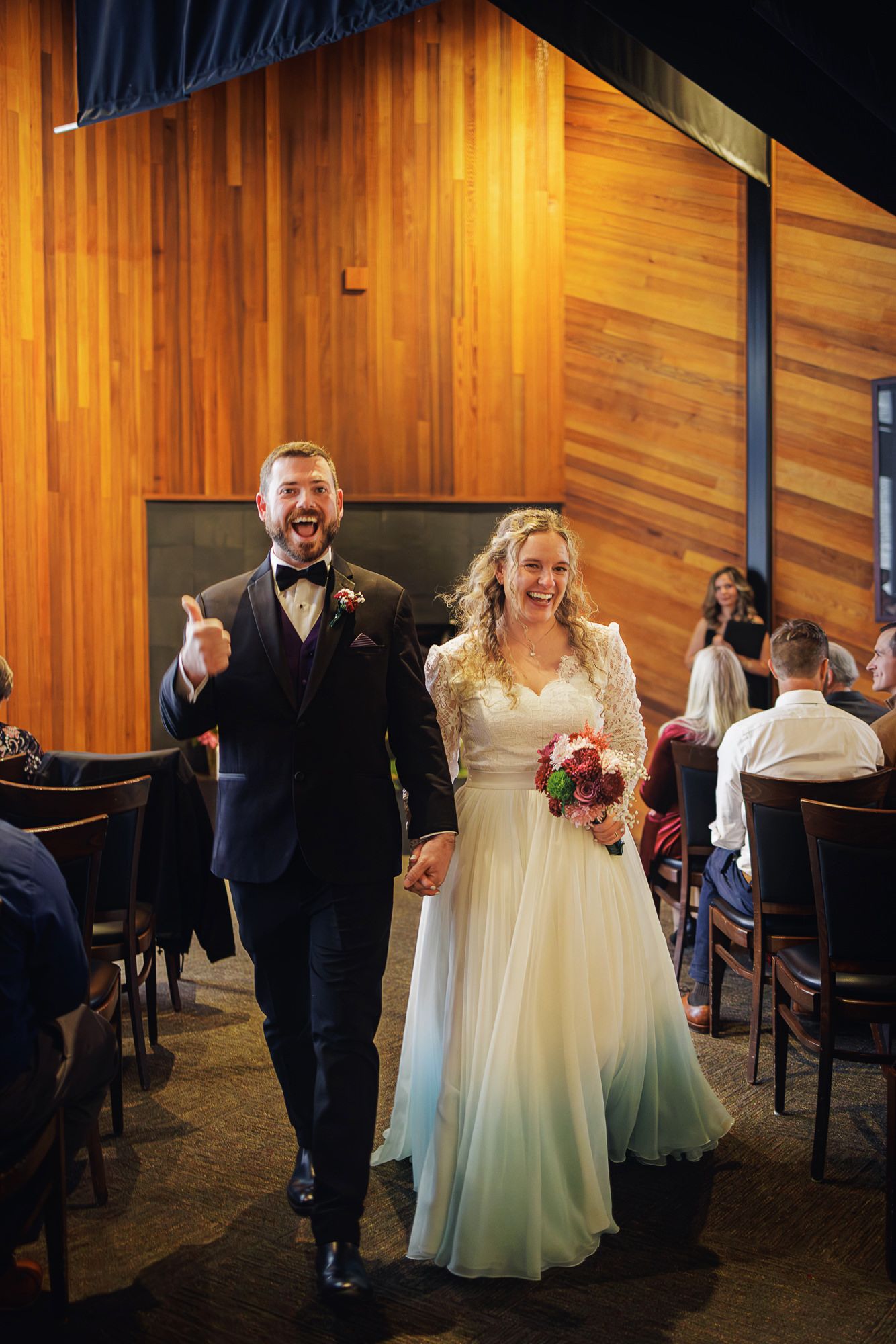 Wedding couple and guests dance joyfully in a rustic wood-paneled venue with warm lighting.