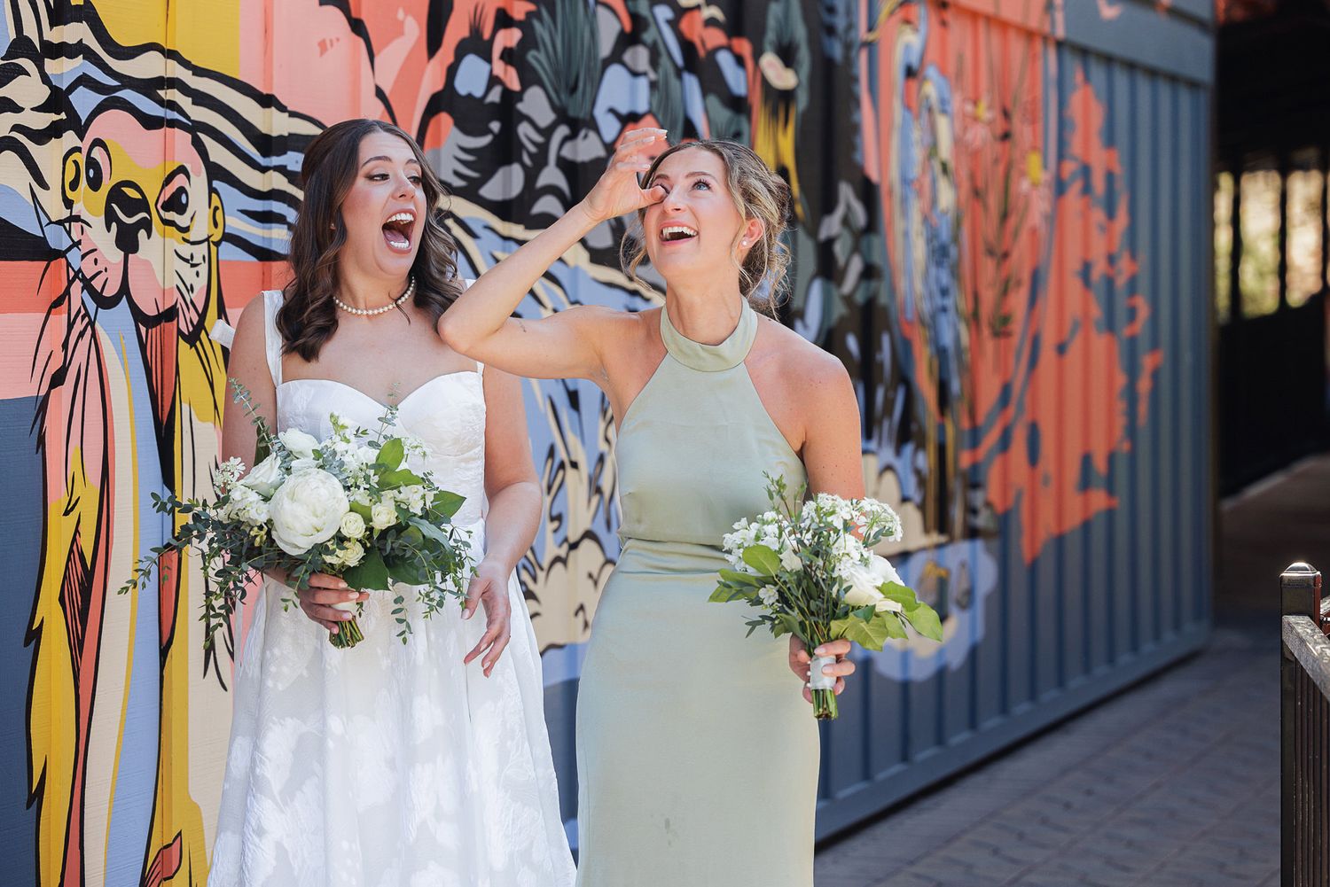 Bride and bridesmaid laughing joyfully holding bouquets in front of a colorful mural.