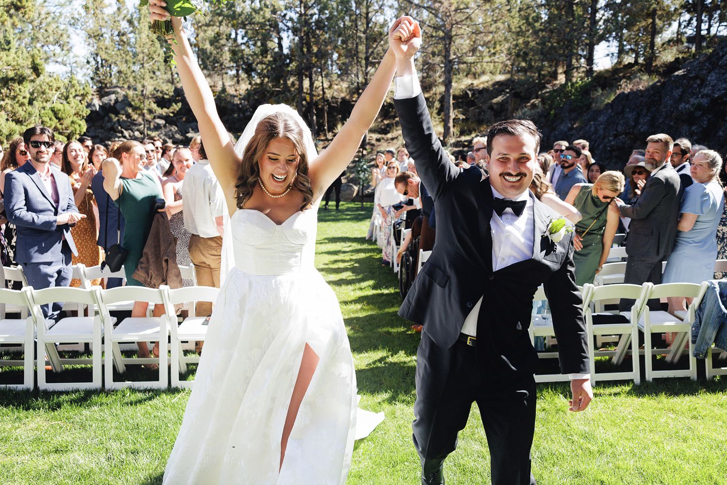 Jubilant newlyweds raise hands in celebration while walking back down the aisle after outdoor ceremony.
