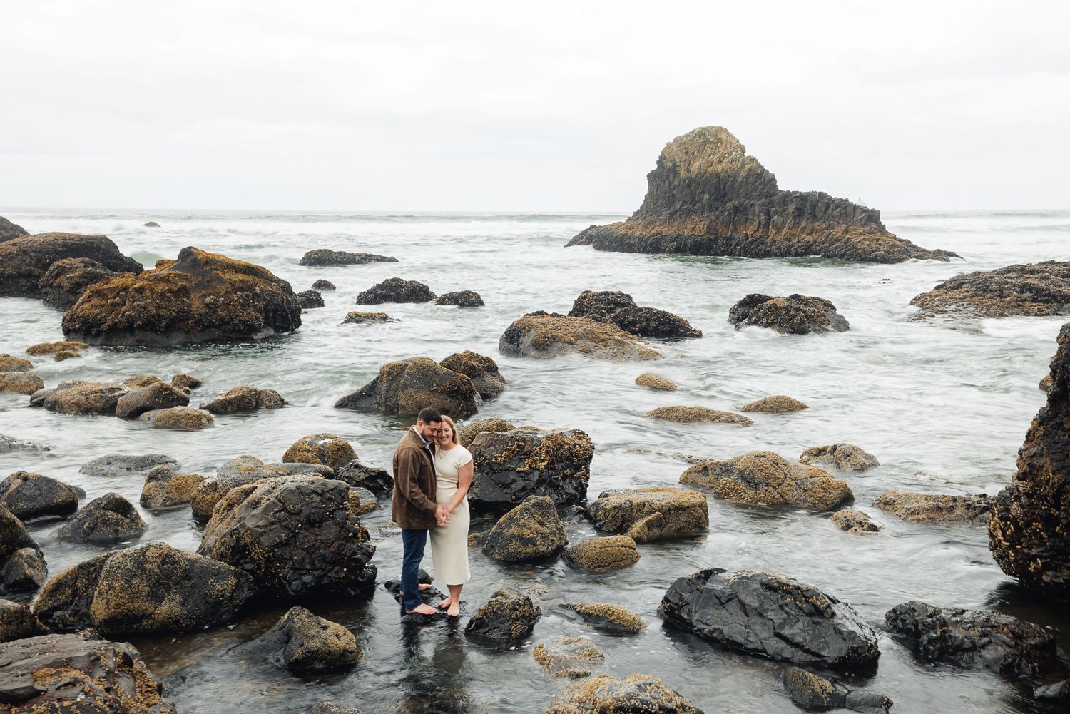Couple embracing among rocky coastal tide pools with misty ocean waves and large boulders on an overcast day.