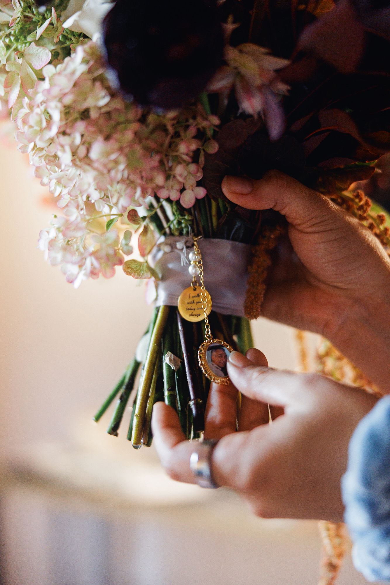 Hands attaching a gold engraved charm and locket to a bridal bouquet of pink hydrangeas and purple flowers.