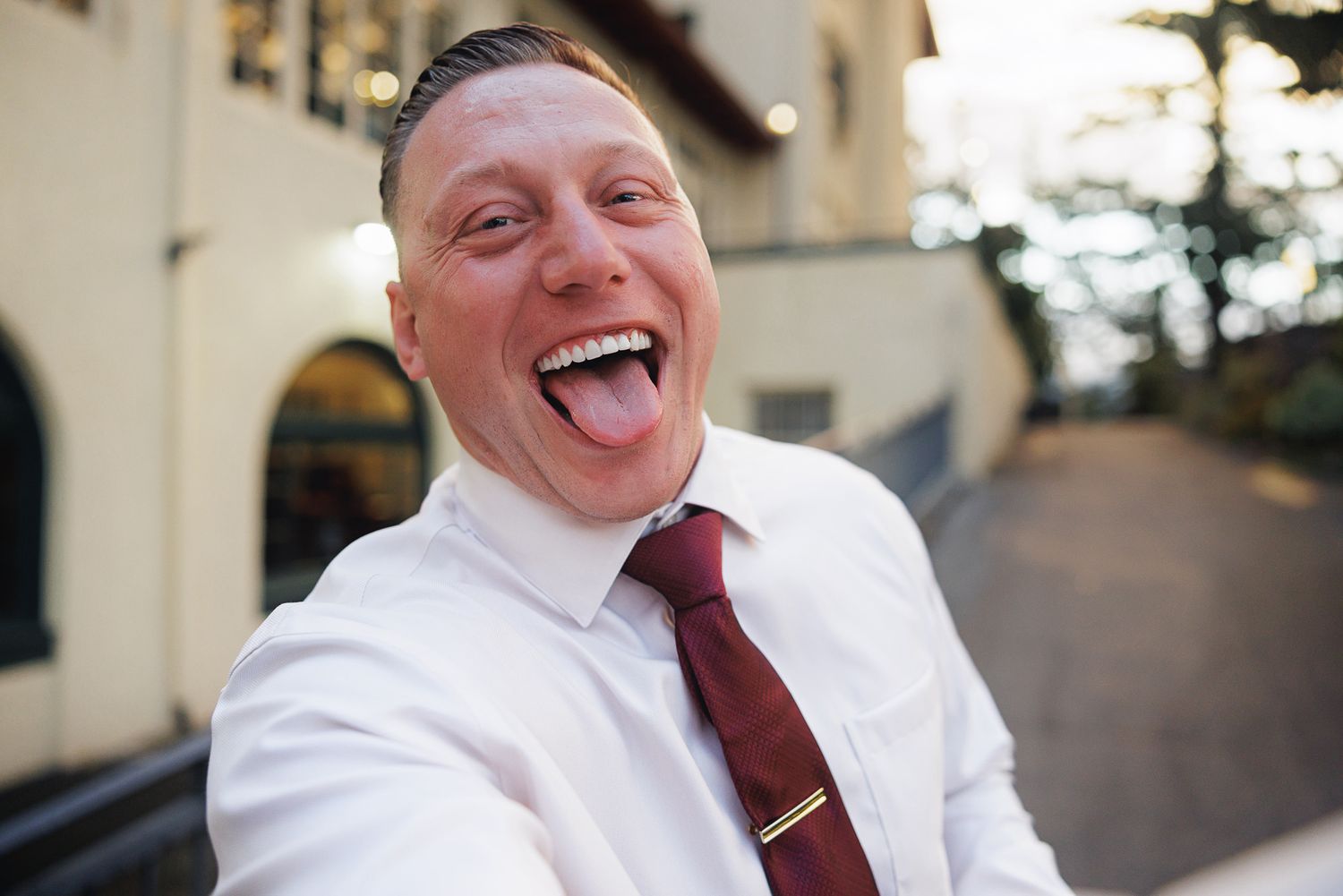 Joyful man in white shirt and burgundy tie takes fun selfie sticking his tongue out at outdoor venue.