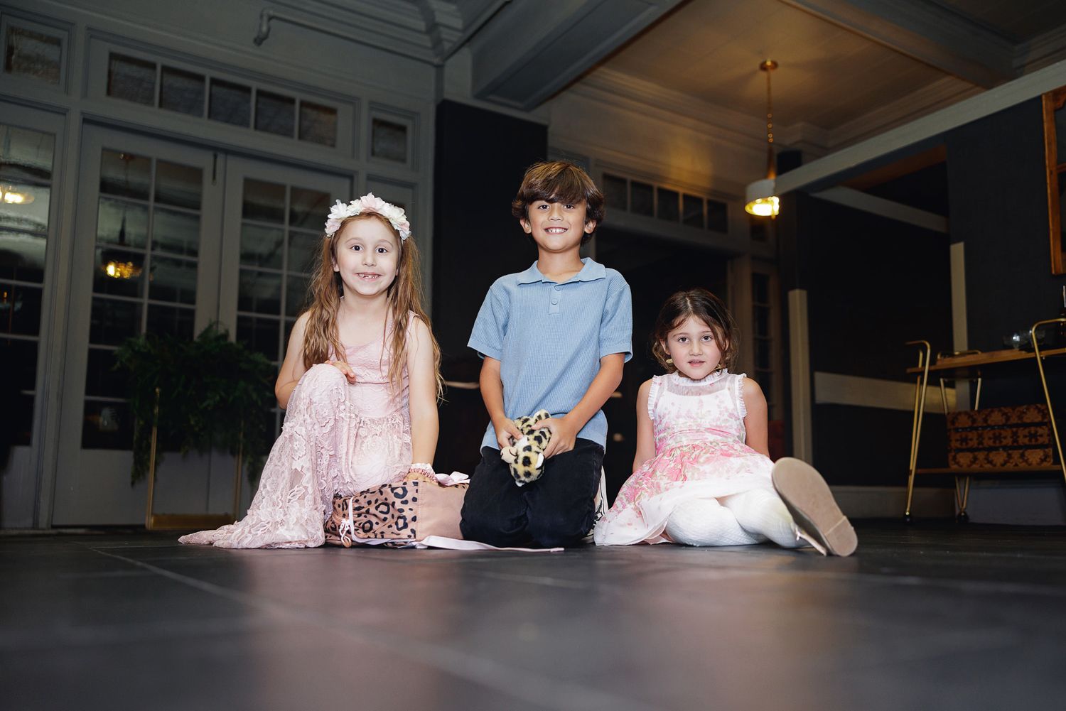 Three children pose on a stage, two girls in pink dresses flanking a boy in a blue shirt during a performance.