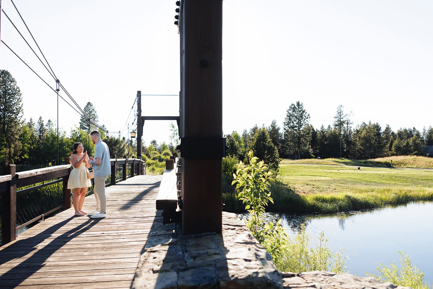 Couple walking on a wooden suspension bridge surrounded by lush greenery and a tranquil pond on a sunny day.