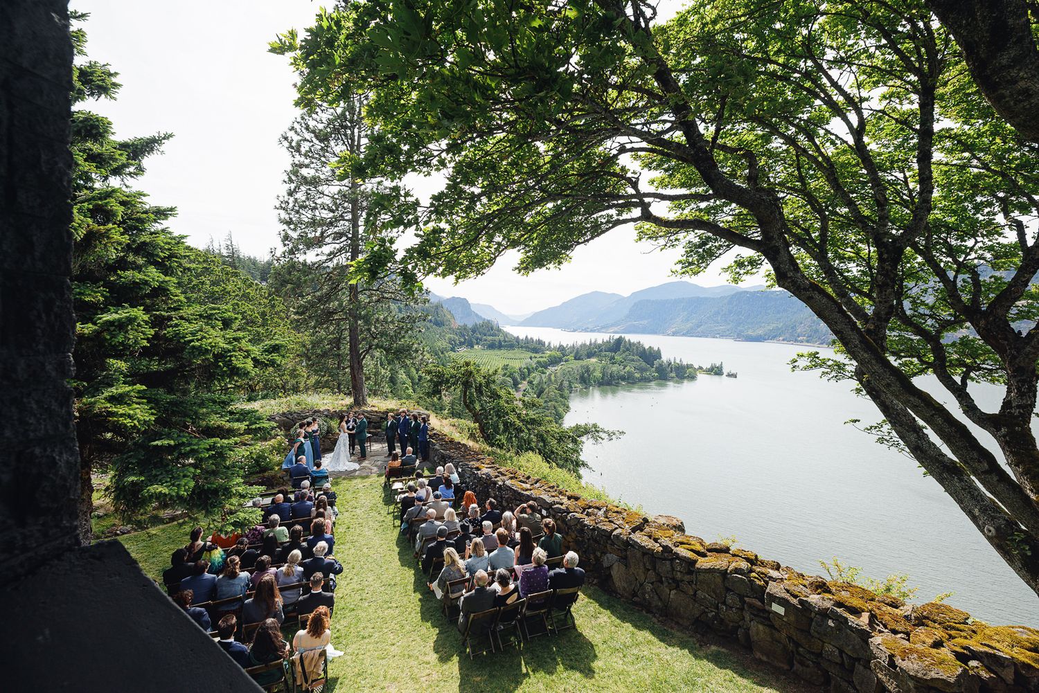Outdoor wedding ceremony on a cliffside terrace overlooking a scenic river and lush green hills under a sunny sky.