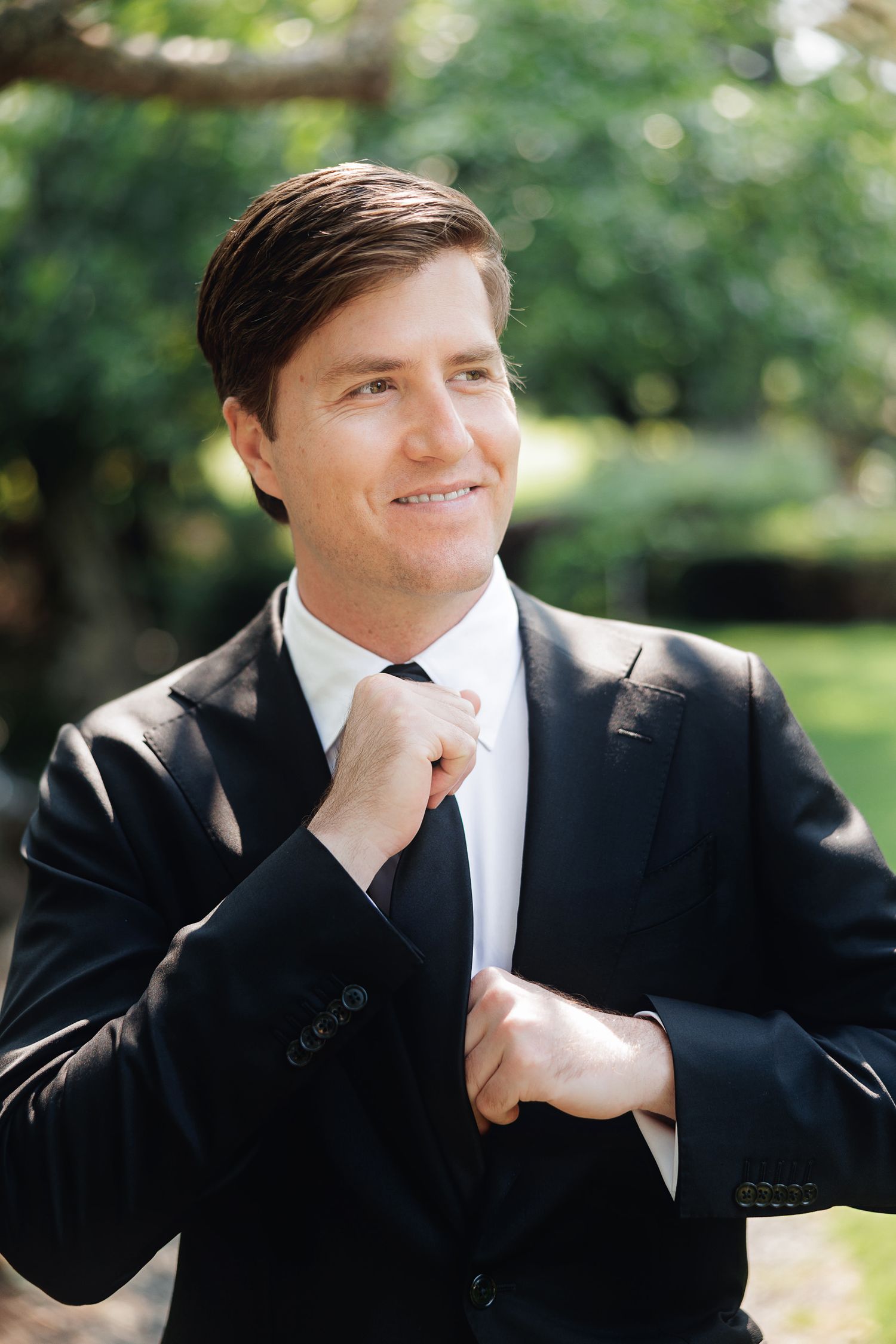 Smiling groom in black suit and tie adjusting jacket outdoors with lush green trees in background.