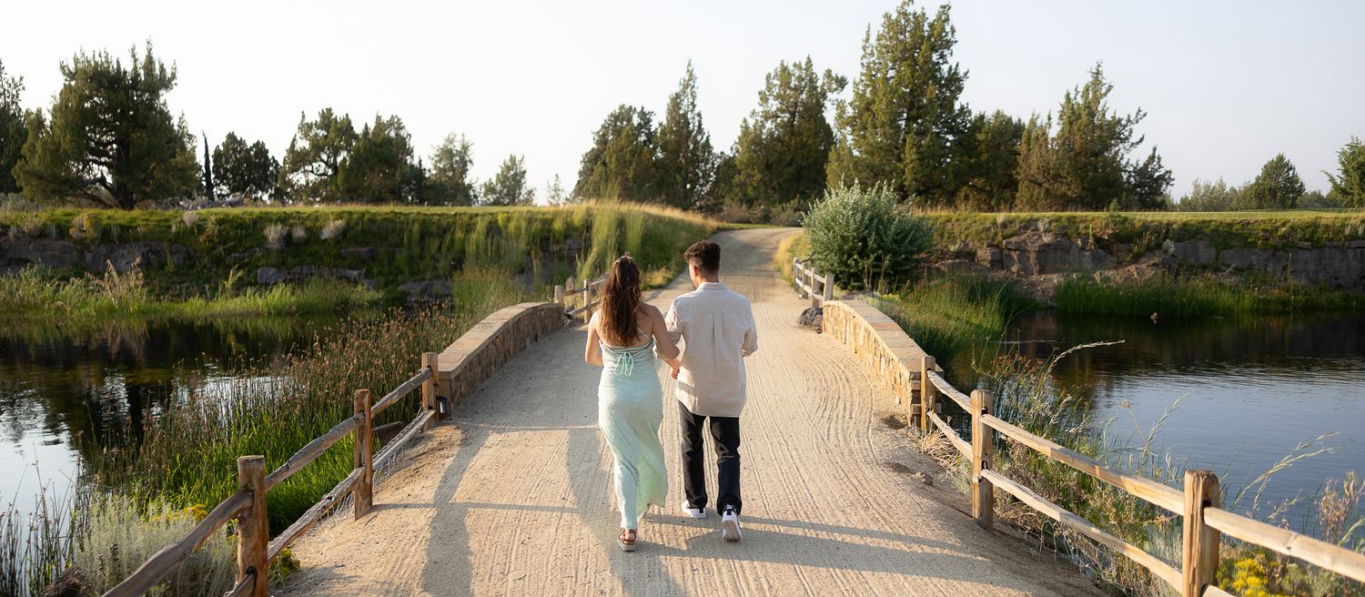 Couple holding hands walking on a wooden bridge surrounded by lush greenery and a serene pond.
