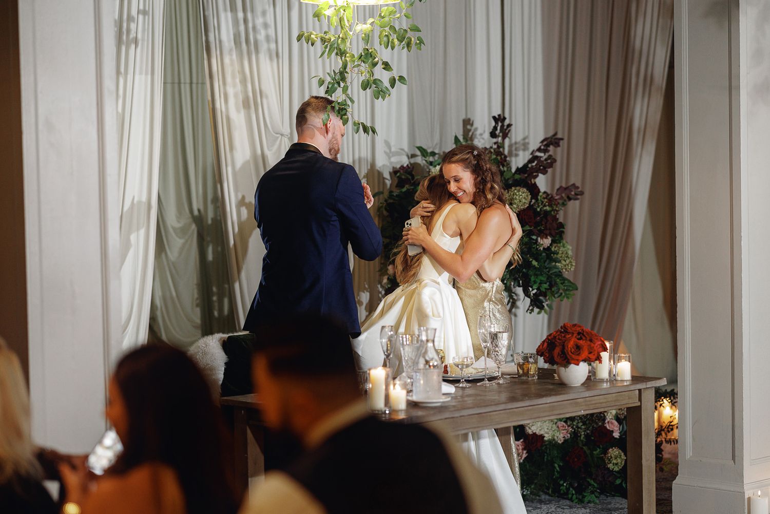 Bride and groom share a joyful embrace at elegant wedding reception with candlelit table and floral arrangements.