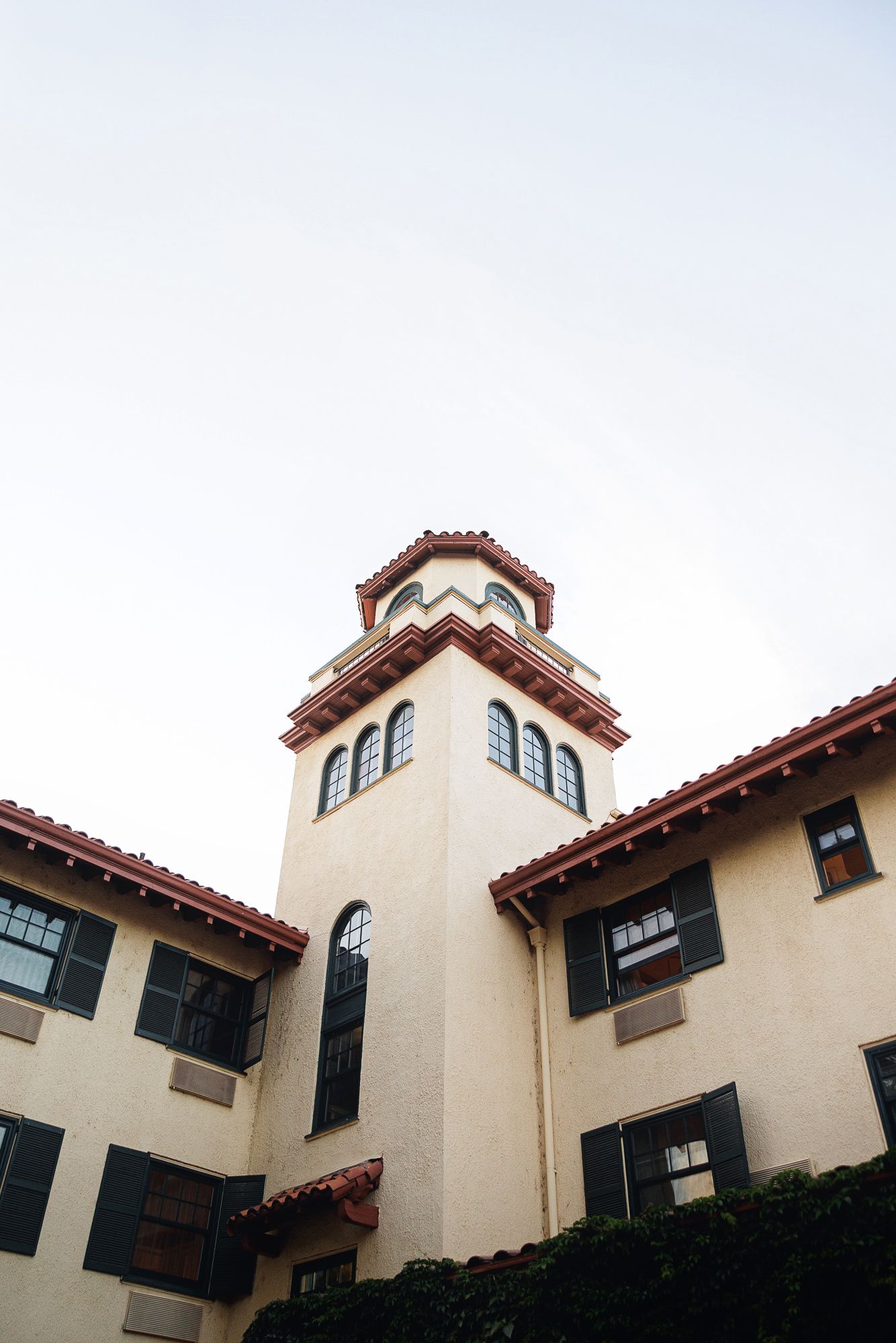 Spanish Colonial Revival building with red tile roof, tower, and black shuttered windows against a pale sky.