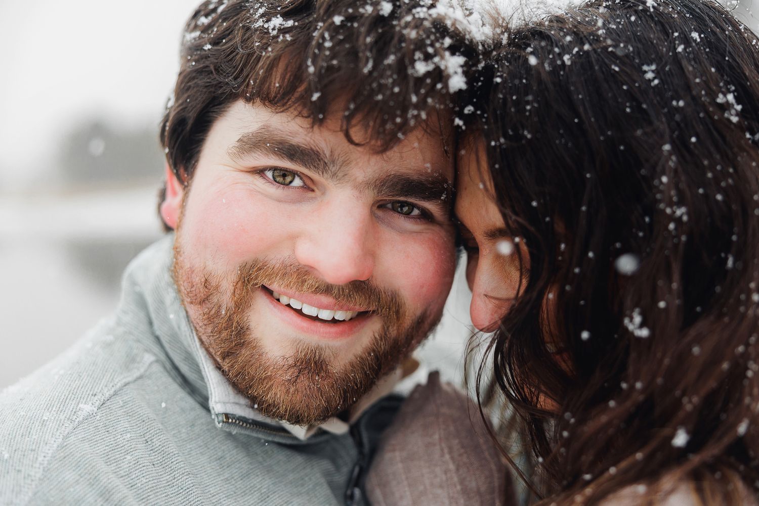 A close-up winter portrait of a couple smiling while snowflakes fall around them.
