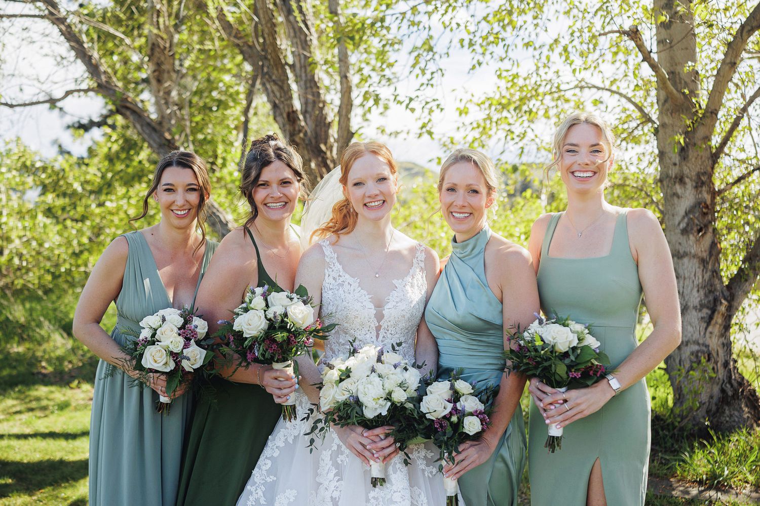 Bride in lace gown with four bridesmaids in sage green dresses holding white bouquets outdoors.