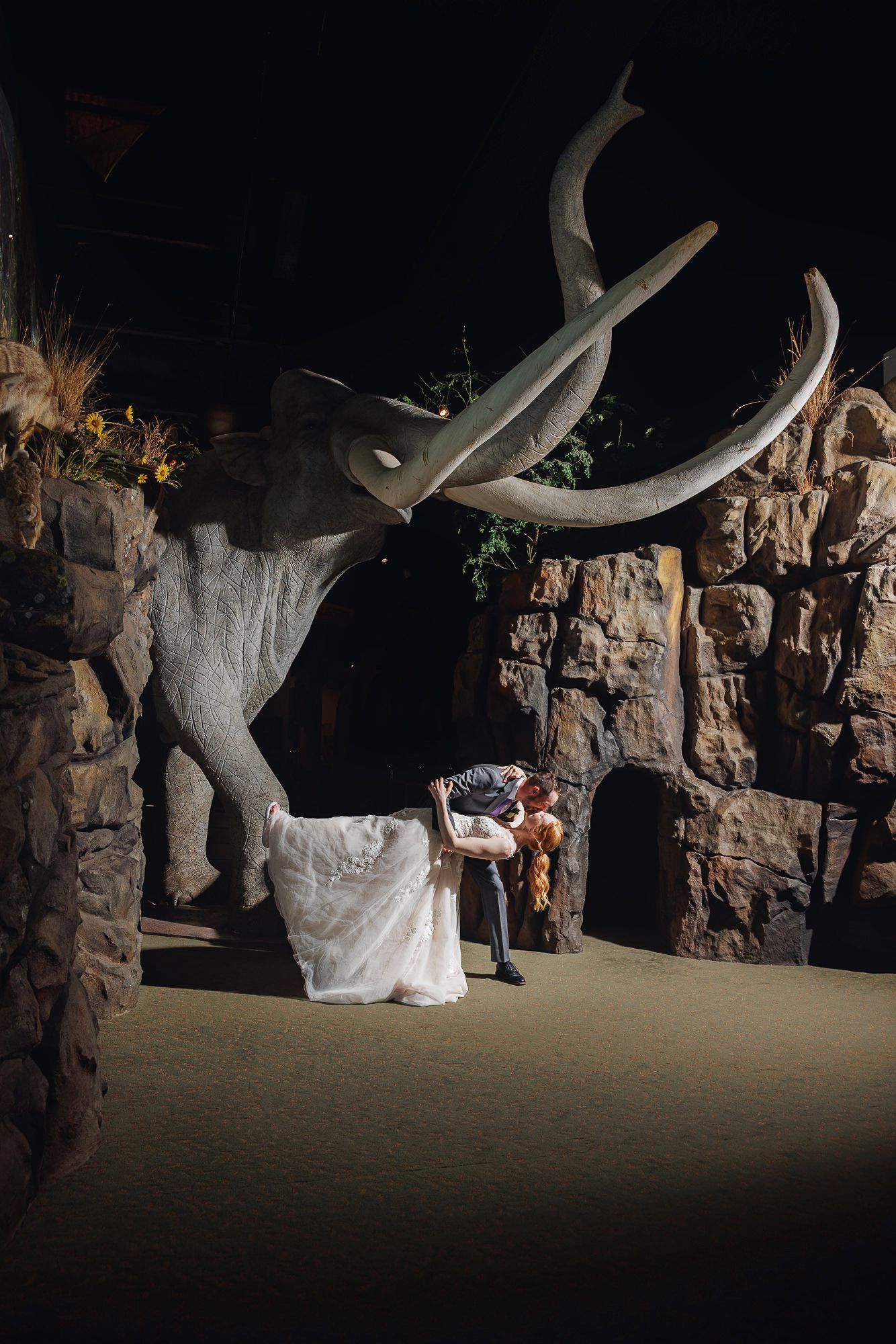 Bride and groom share a romantic dip kiss in front of a large mammoth statue in a dimly lit museum venue.
