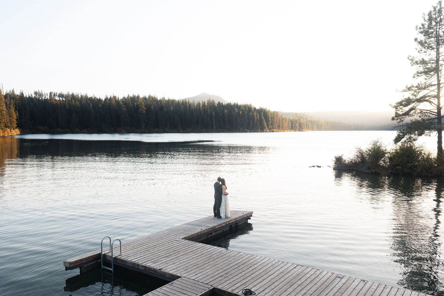 Couple embracing on a wooden dock at sunset over a calm mountain lake surrounded by evergreen forest.