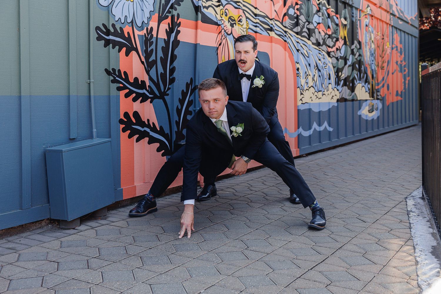 Two grooms in black tuxedos pose playfully in front of a colorful floral mural at their wedding.