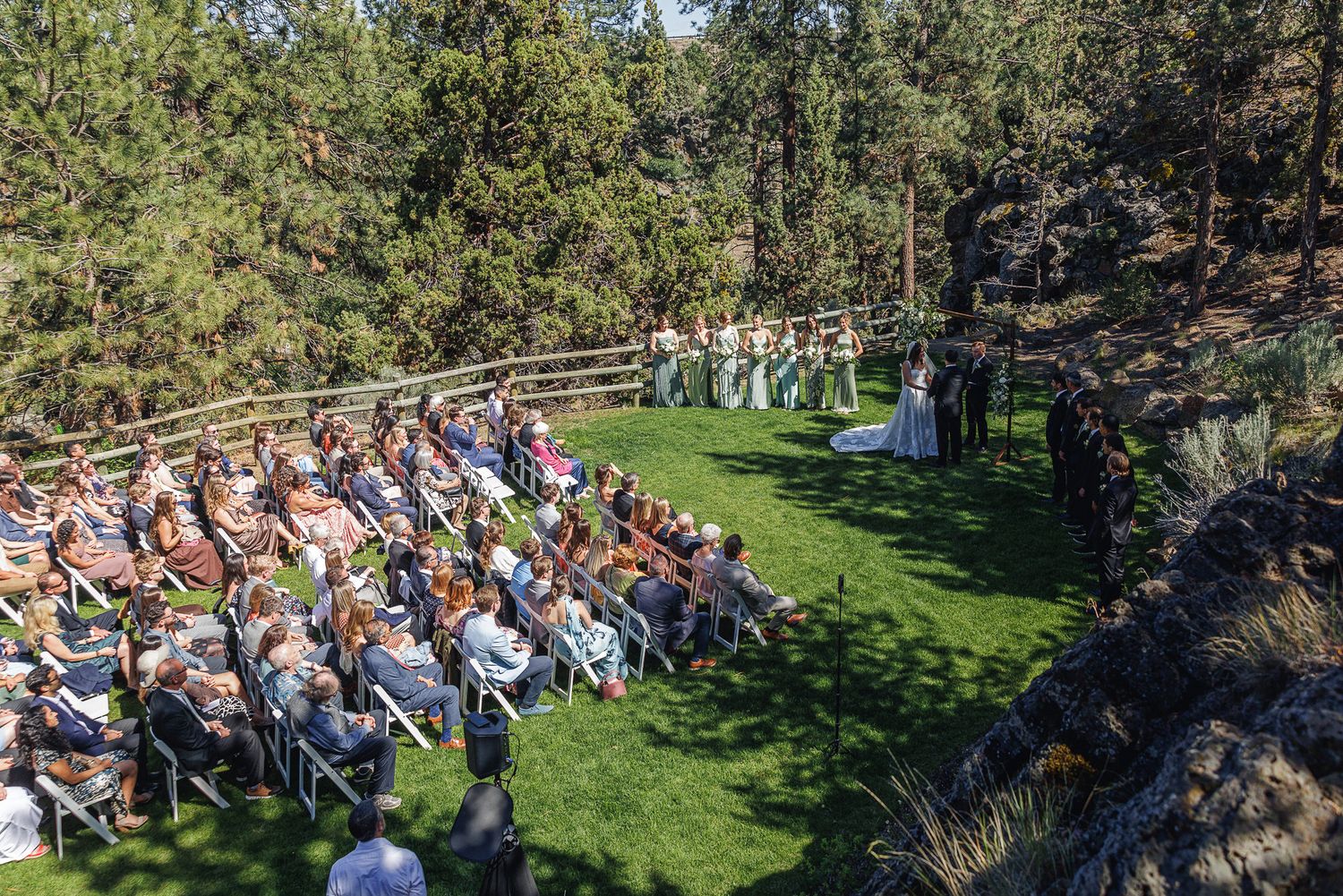 Aerial view of outdoor wedding ceremony with guests seated on white chairs facing altar amid pine trees.