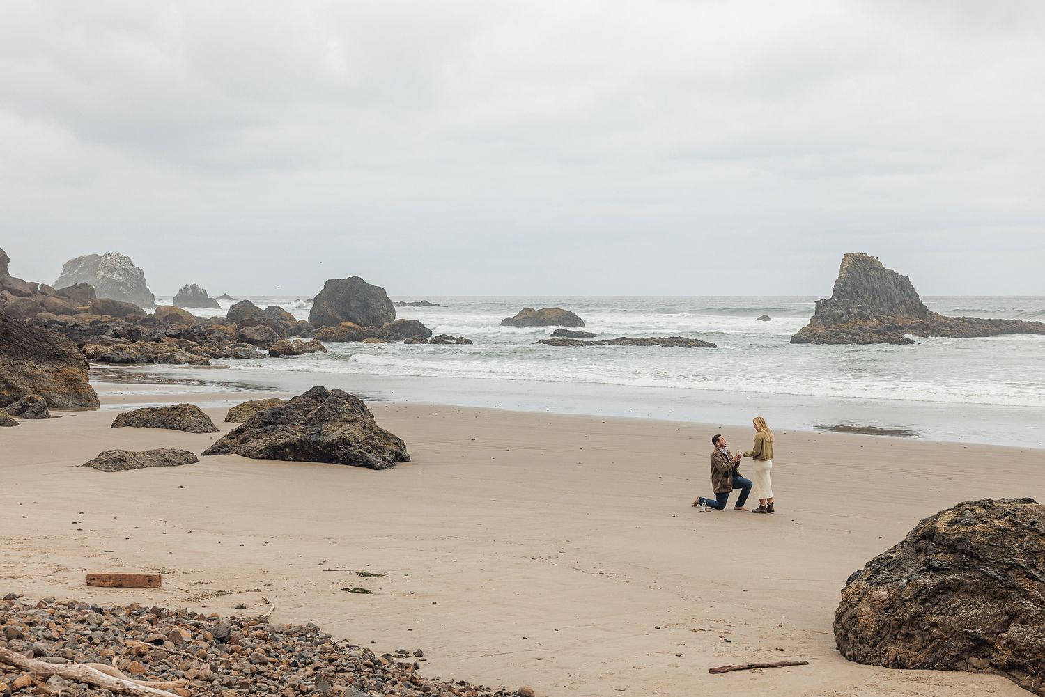 Couple on a sandy beach with rocky sea stacks and ocean waves under an overcast sky.