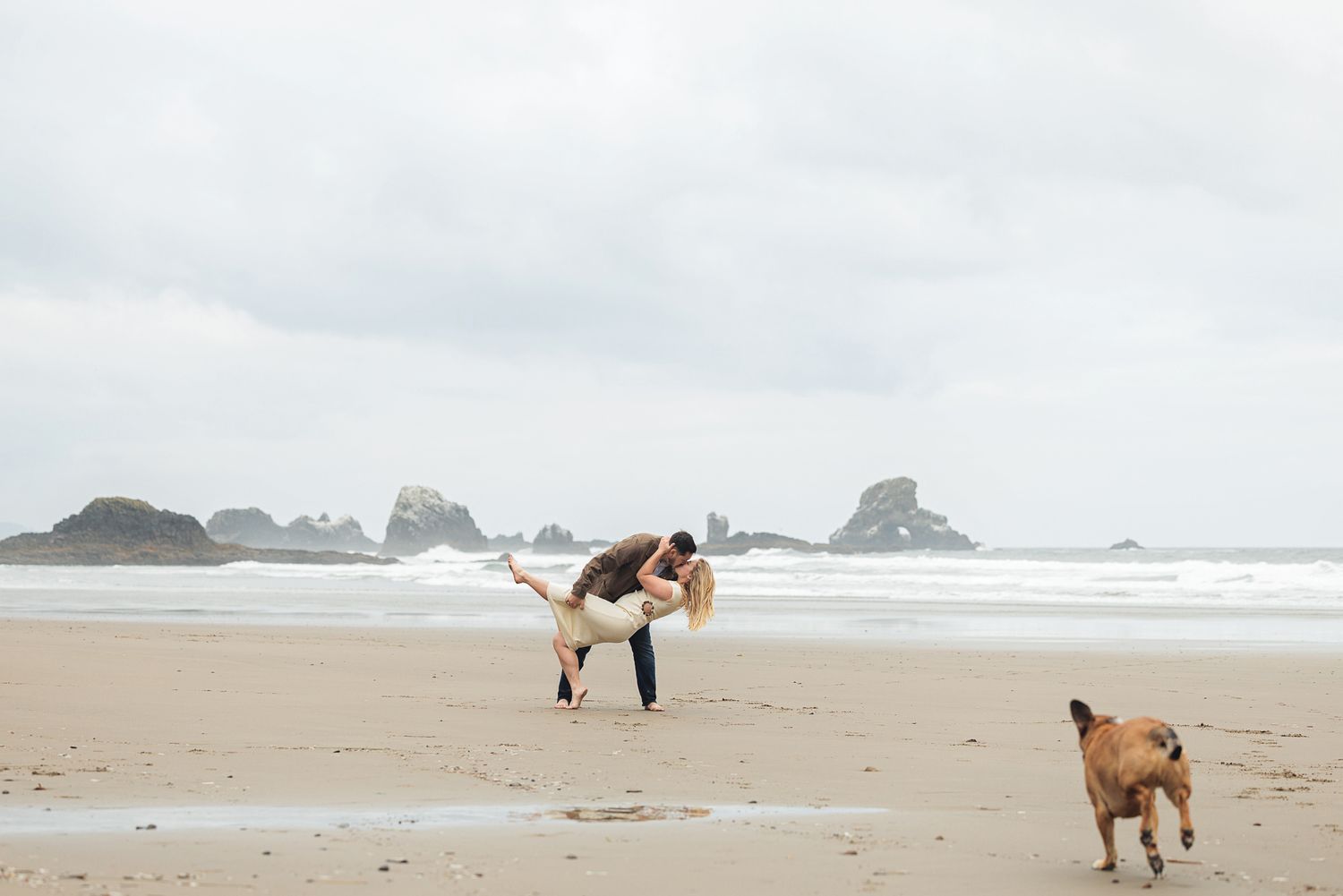 Couple dancing romantically on a sandy beach with a dog nearby and rocky ocean formations in the background.