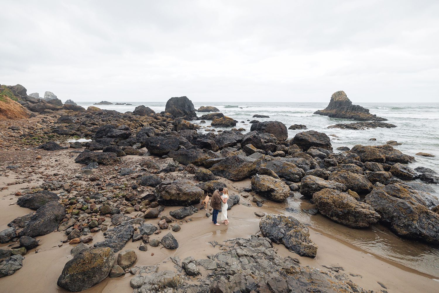Couple standing among rugged coastal rocks on a sandy beach with ocean waves and misty sky.
