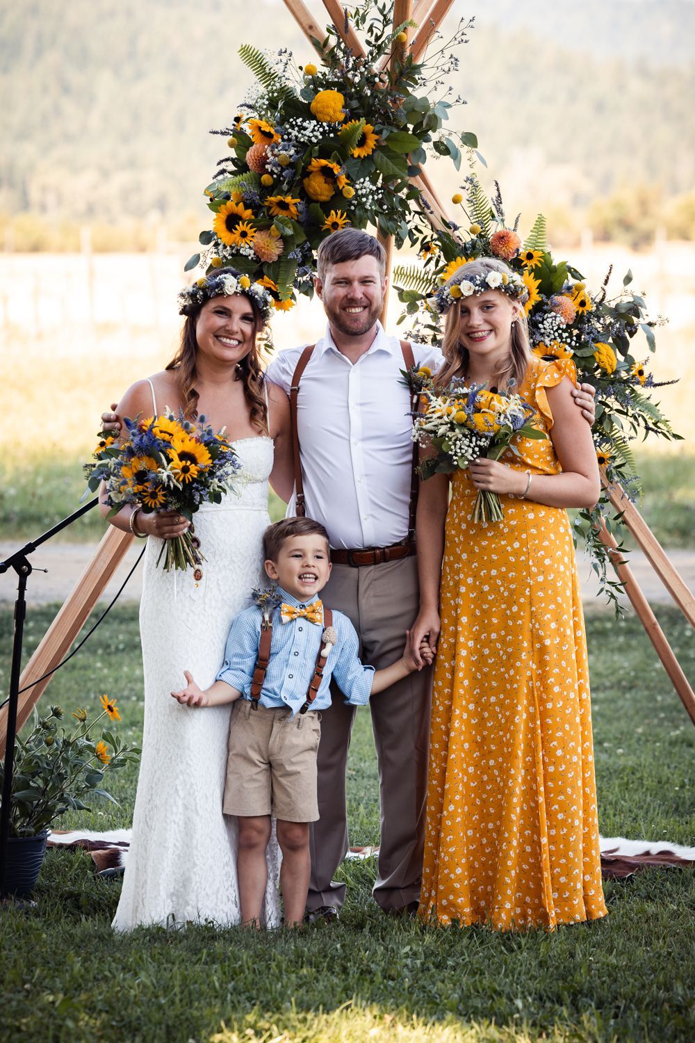 Outdoor wedding party standing under floral arch with yellow sunflowers and blue flowers on rustic wooden frame in field.