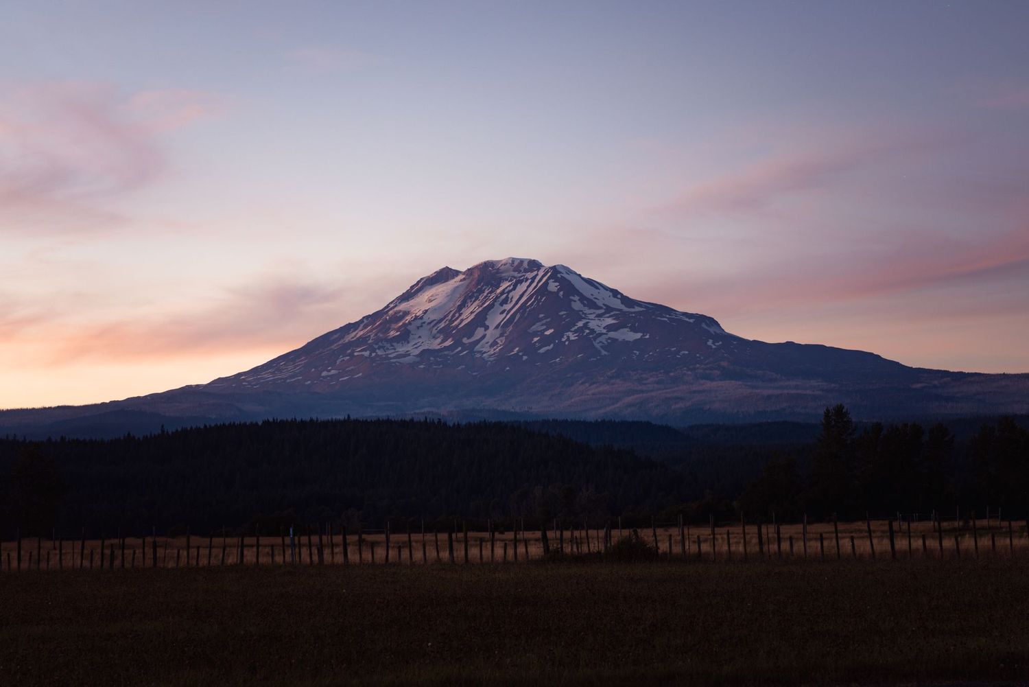 Snow-capped Mount Adams rises against a pink sunset sky with a wooden fence in the foreground in Washington state.