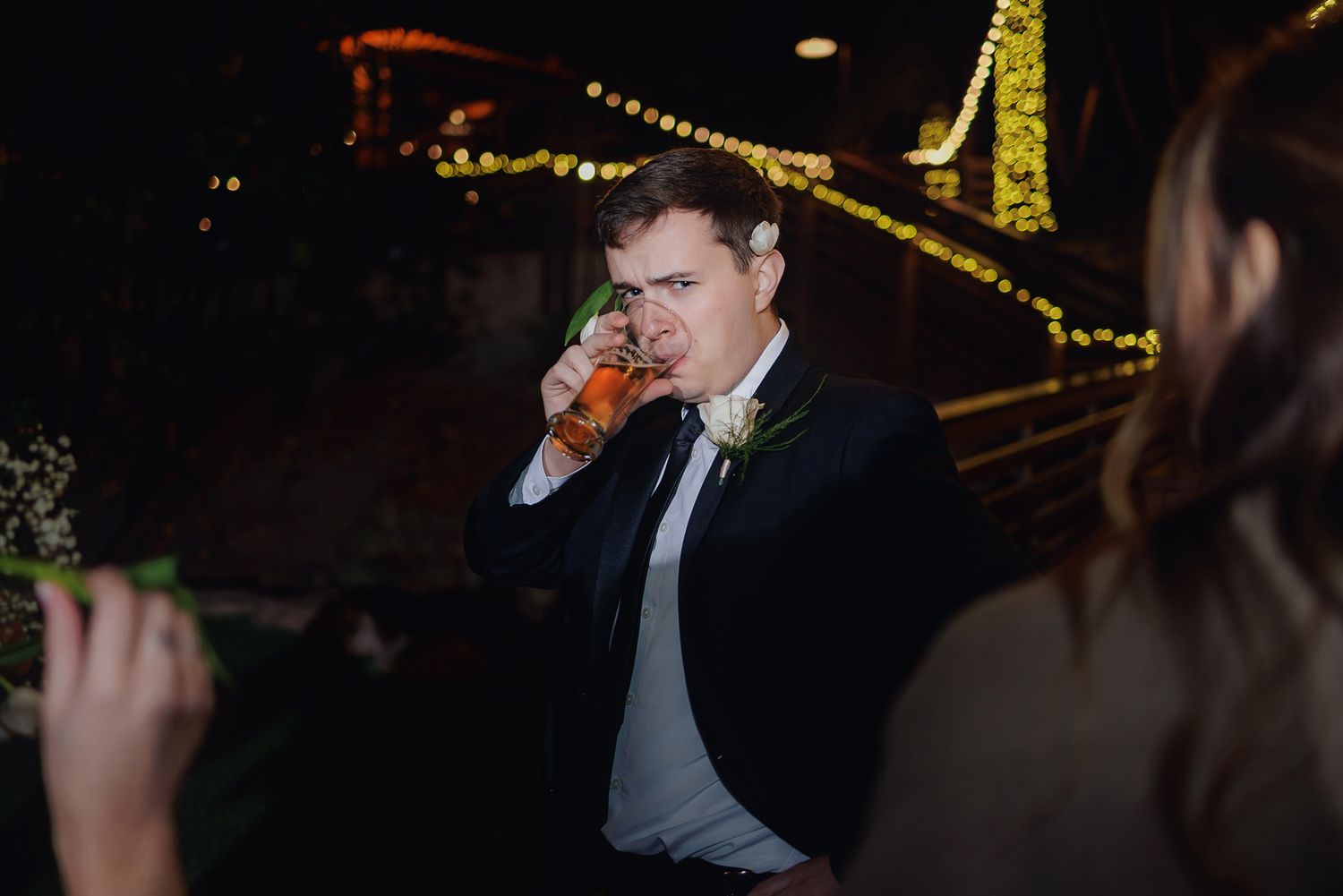 A person in formal attire sips a beverage at an evening event with twinkle lights visible in the background.