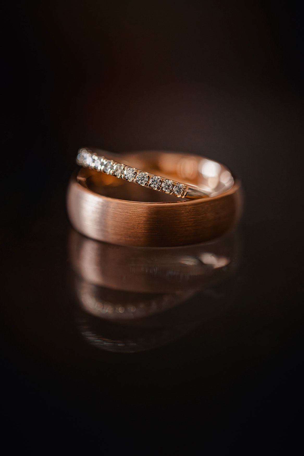 Two wedding rings positioned together on dark surface, one brushed rose gold band and one diamond-encrusted band.