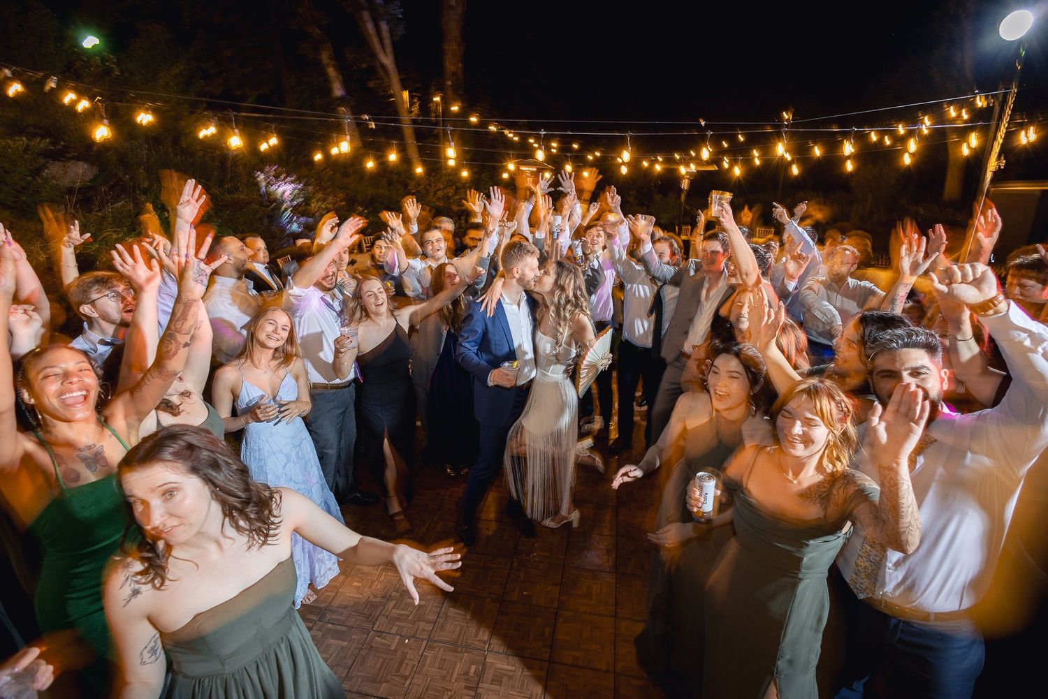 Wedding guests dance and celebrate under glowing string lights at an outdoor evening reception party.