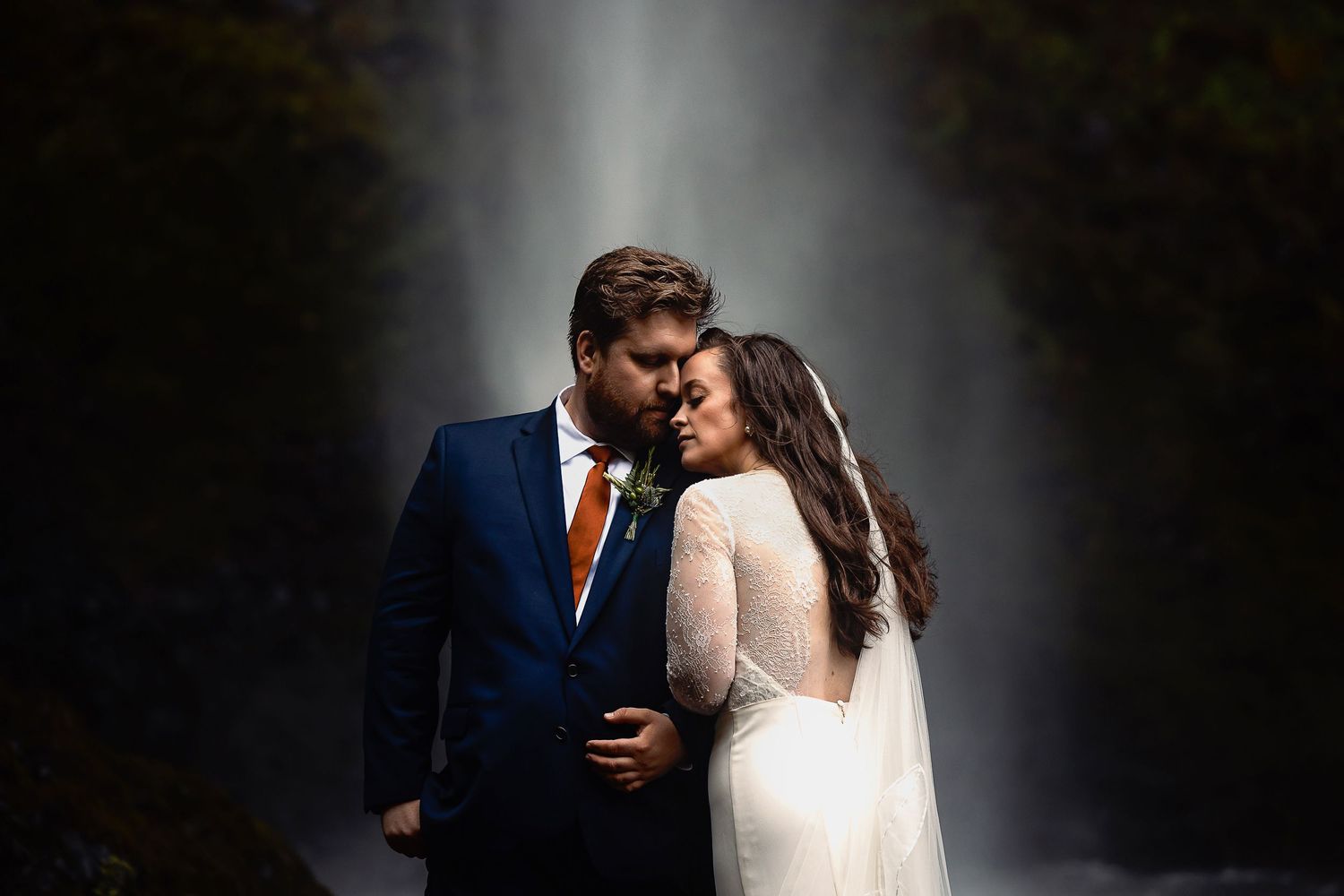 Dramatic wedding portrait with waterfall backdrop creating ethereal lighting effect.
