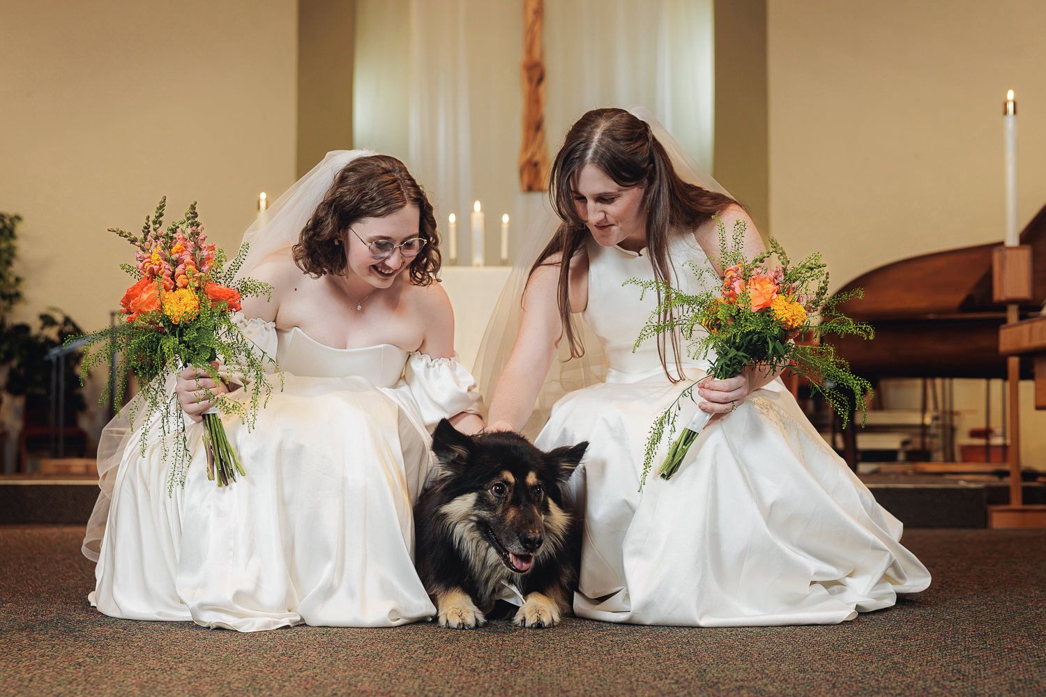 Two people in white wedding dresses sit on the floor petting a black and brown dog during a photo session.