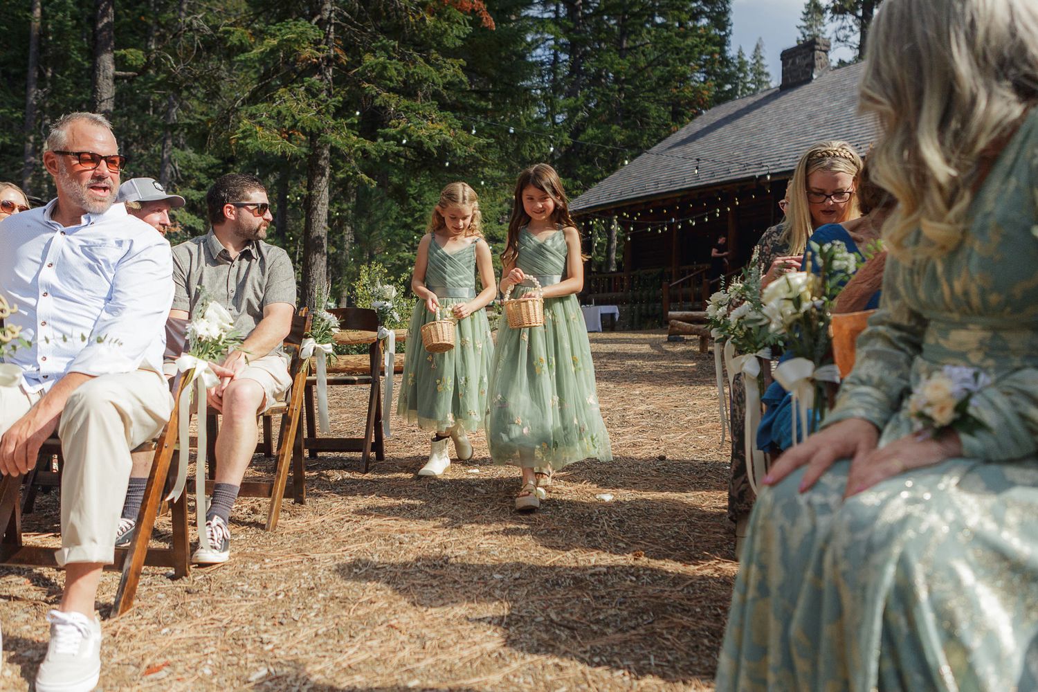 Wedding party celebrates outdoors in a wooded setting with sage green bridesmaid dresses and string lights overhead.