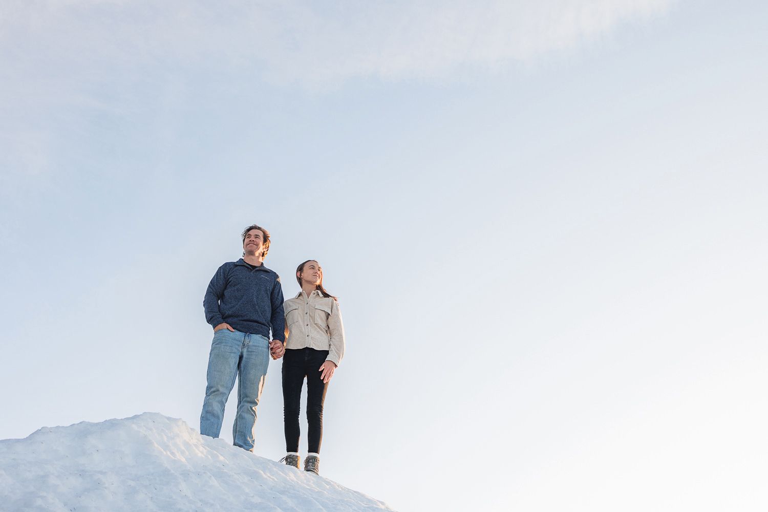 Two people stand atop a snowy hill against a light blue sky on a winter day.