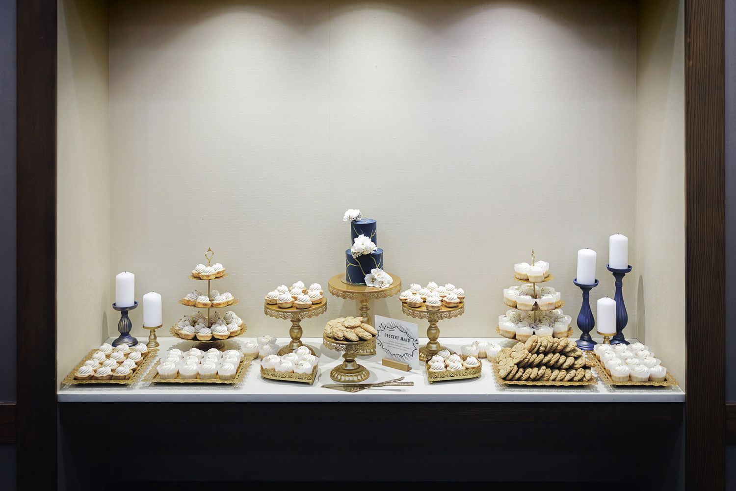 An elegant dessert display featuring white cupcakes and treats arranged on gold stands with navy blue accents.