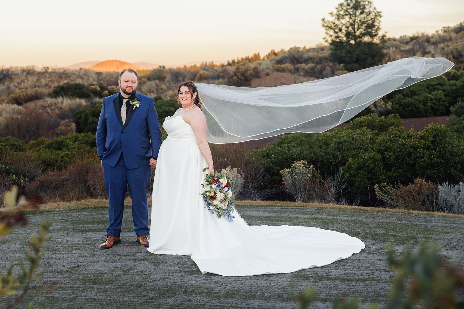A couple in wedding attire poses outdoors at sunset with a flowing veil caught in the wind.
