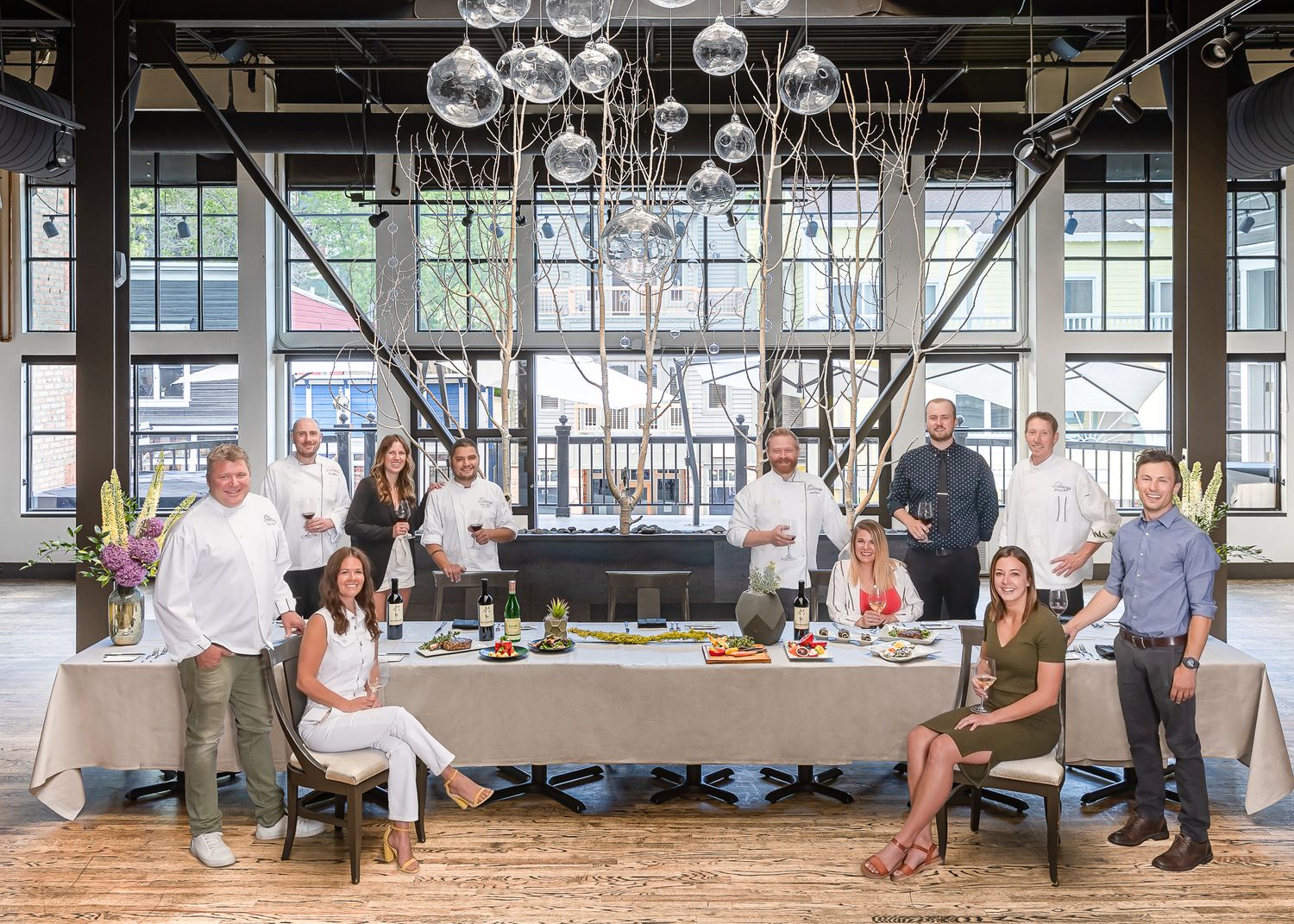 Elegant indoor gathering with long dining table, hanging light fixtures and industrial windows.