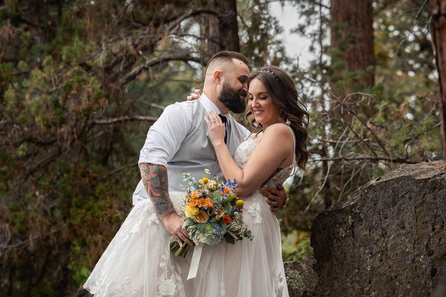 Two people share a tender moment in wedding attire surrounded by pine trees.