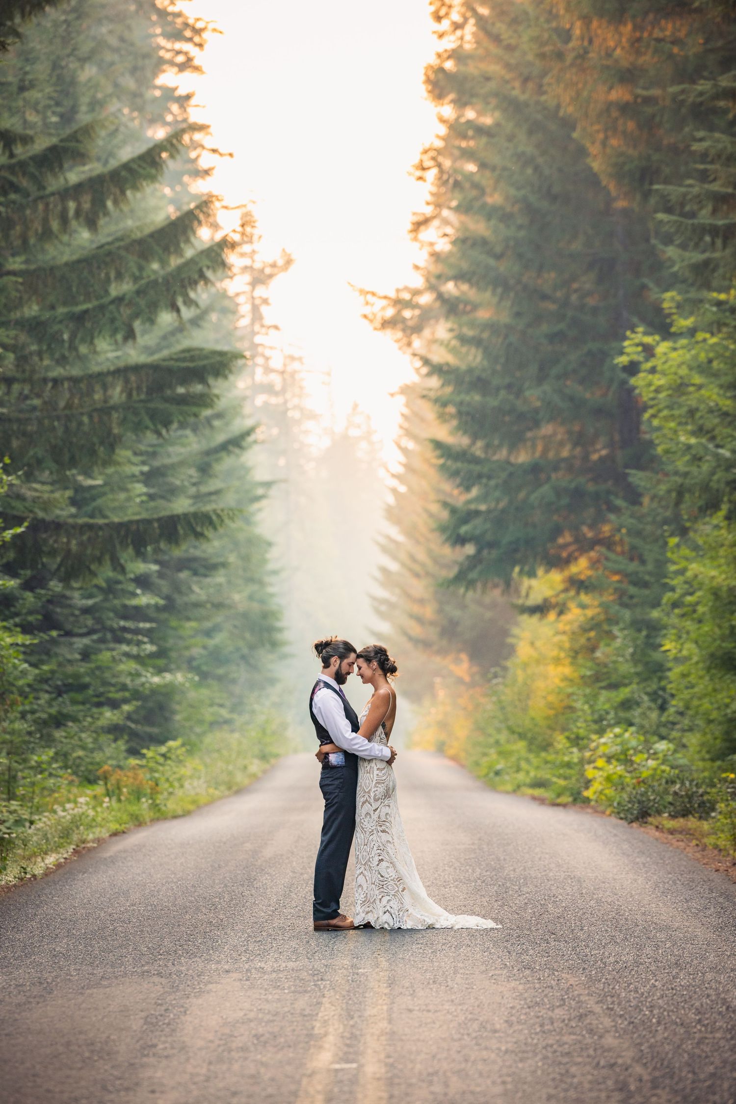 Romantic forest road wedding portrait with tall evergreen trees and golden light.
